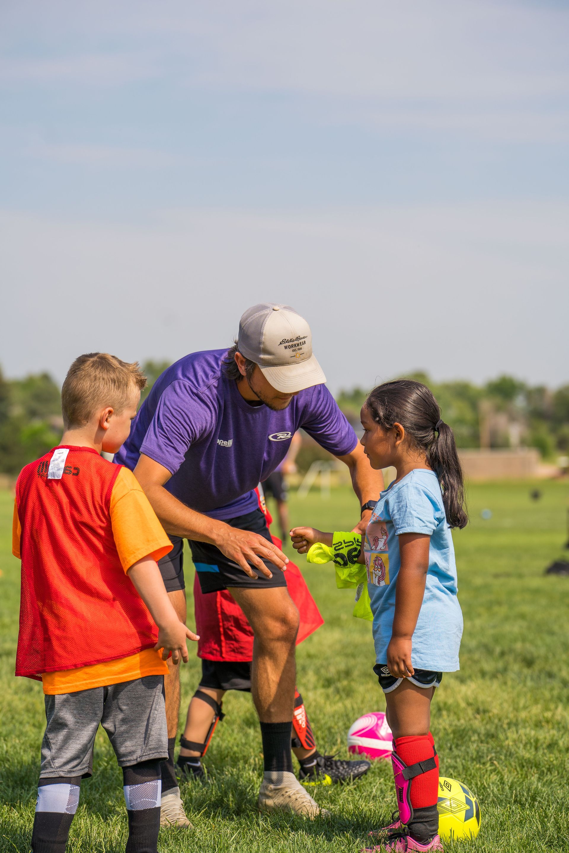 Coach instructing two young soccer players on a field, with cones and balls nearby