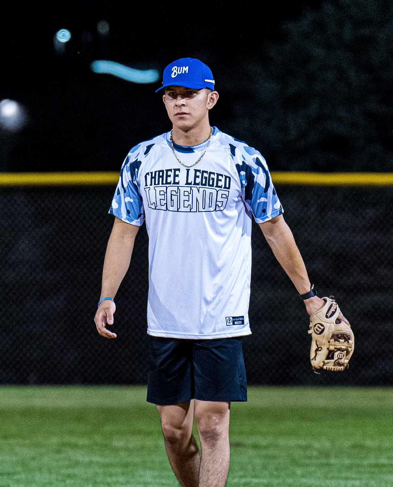 Baseball player in white jersey and blue cap standing on a lit field at night, holding a glove.