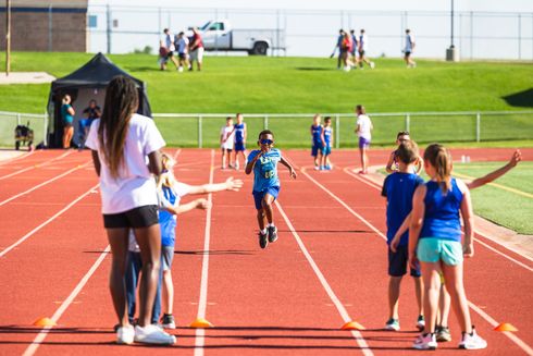 Track meet with children running on a red track, others standing nearby, and a grassy field in the background