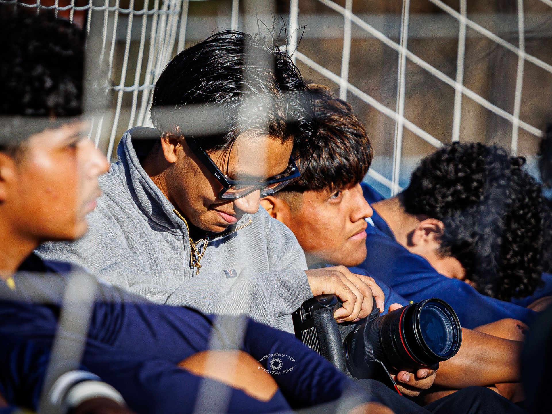 Several people sit behind a chain-link fence, one holding a professional camera with a large lens.