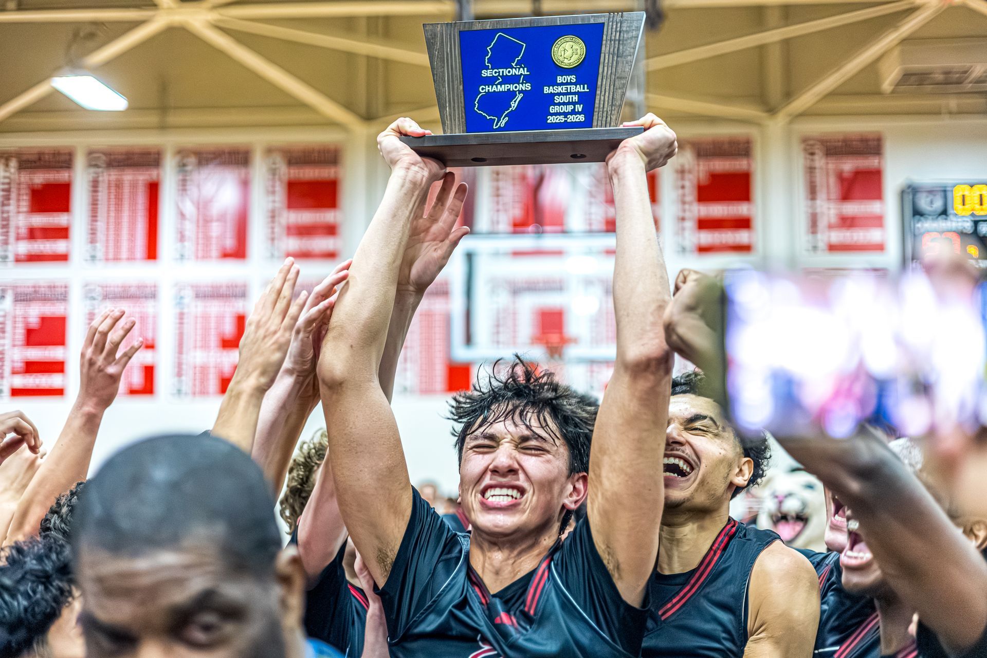 Athletes in dark uniforms joyfully lift a championship trophy high above their heads in a basketball gymnasium.