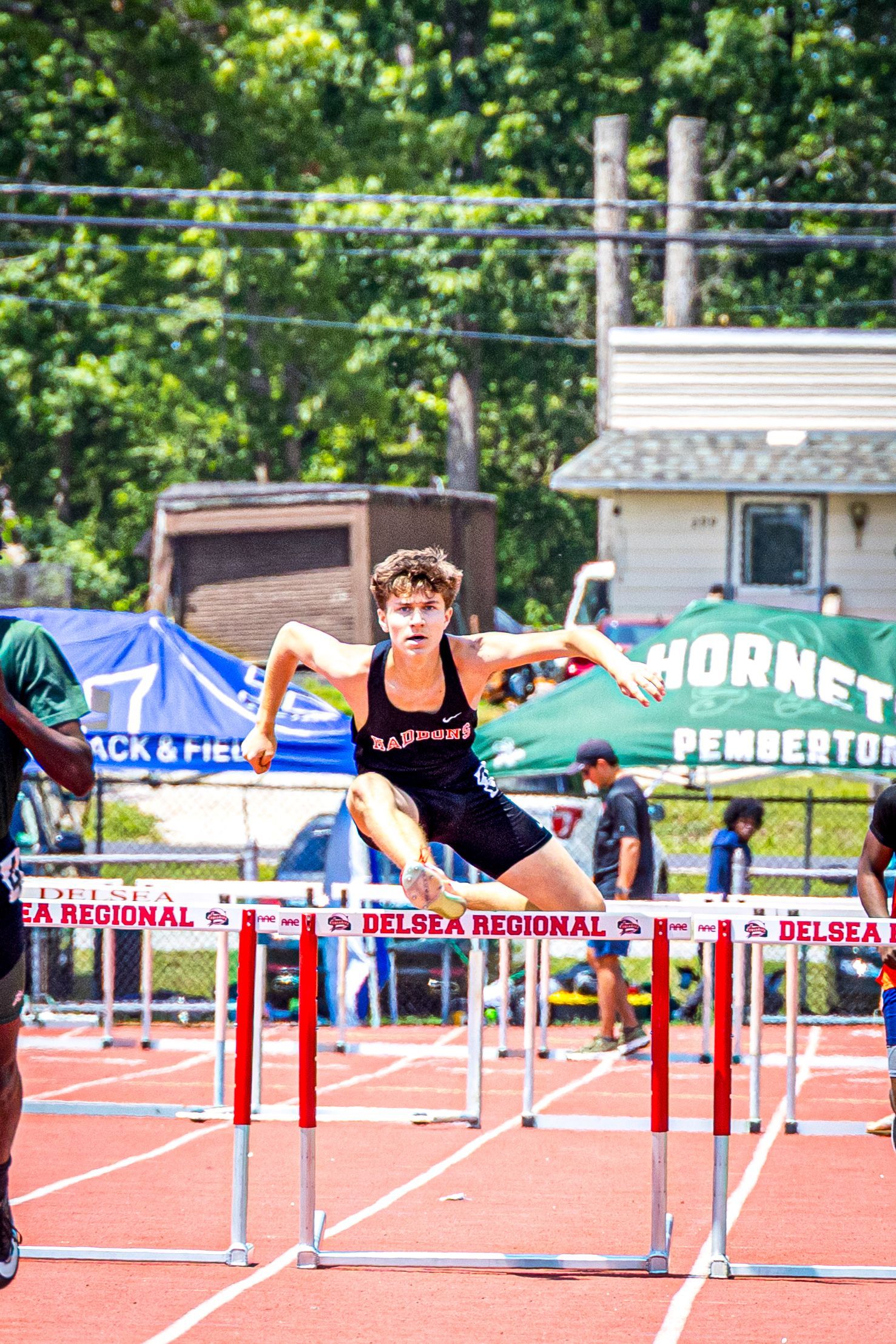 A runner in a black track uniform clears a hurdle on an outdoor track during a race.