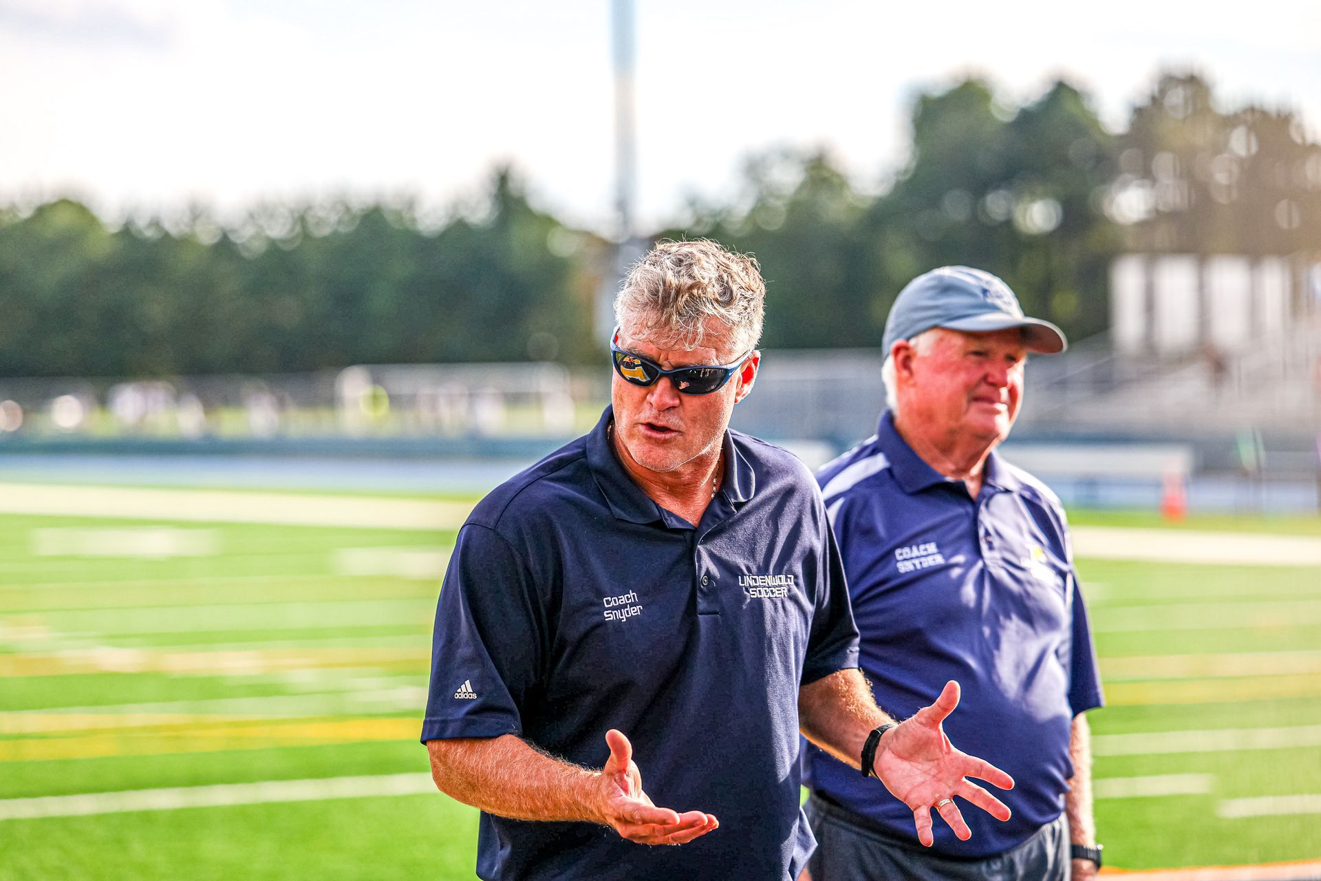 Two men in athletic polos stand on a green football field, with one speaking while gesturing with his hands.