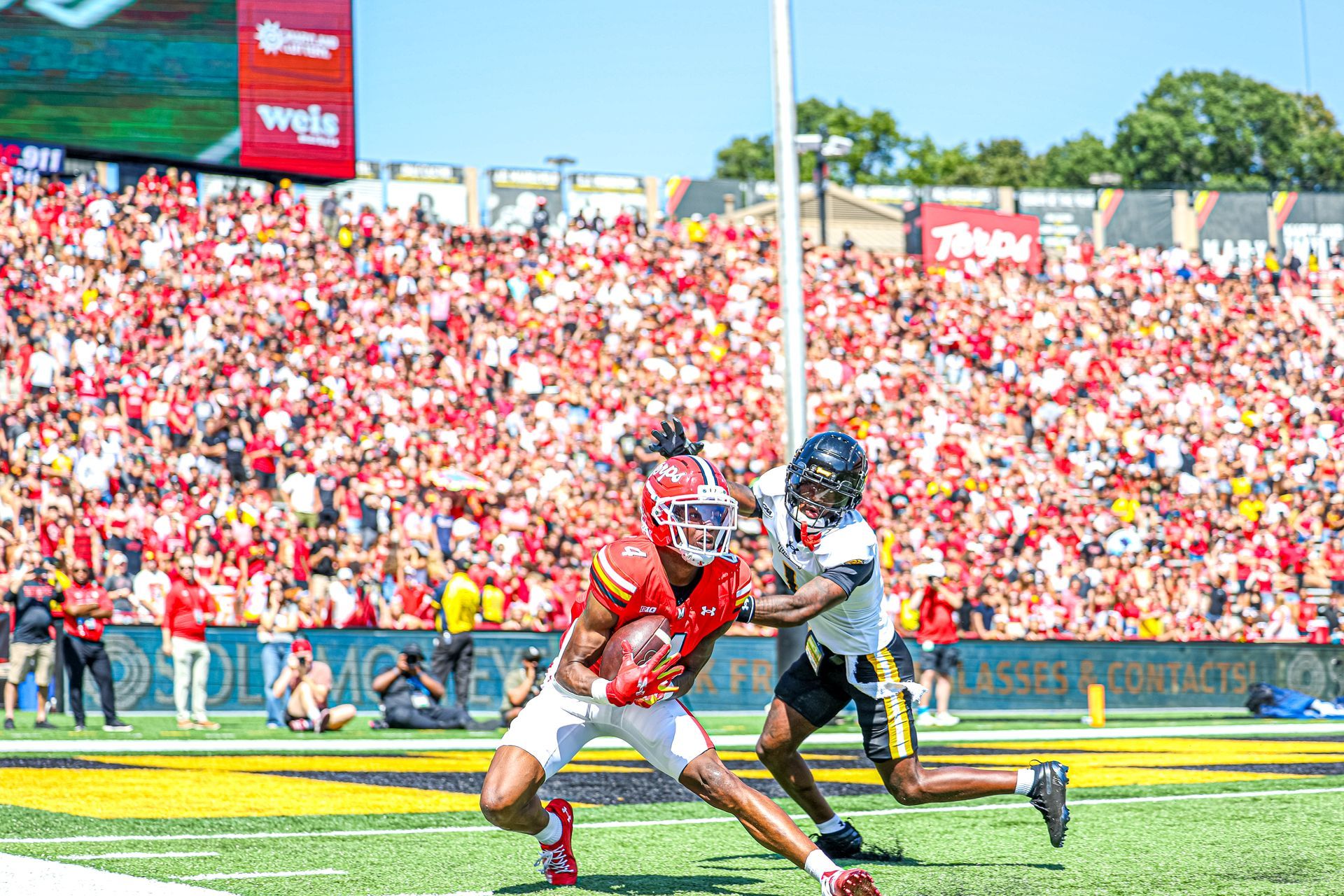 A college football player in a red uniform carries the ball past a defender in white during a game in a crowded stadium.
