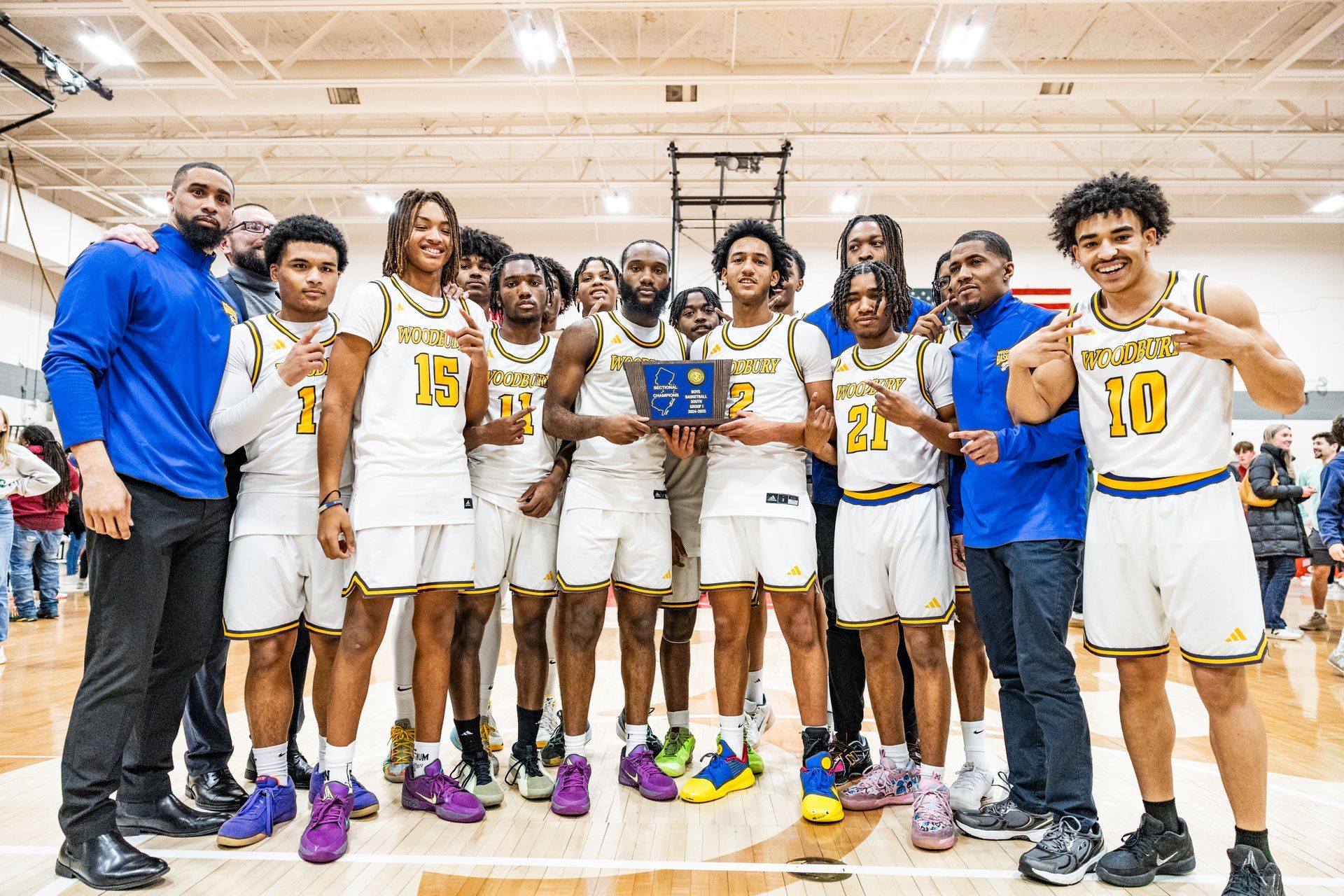 A basketball team in white and blue uniforms poses with a championship trophy in a brightly lit gymnasium.
