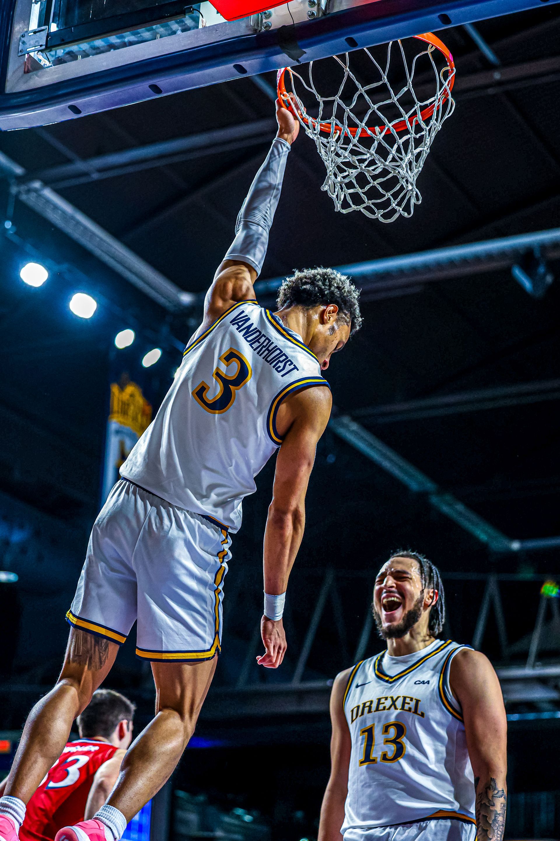 A basketball player in a white uniform dunks a ball while a teammate celebrates below on the court.
