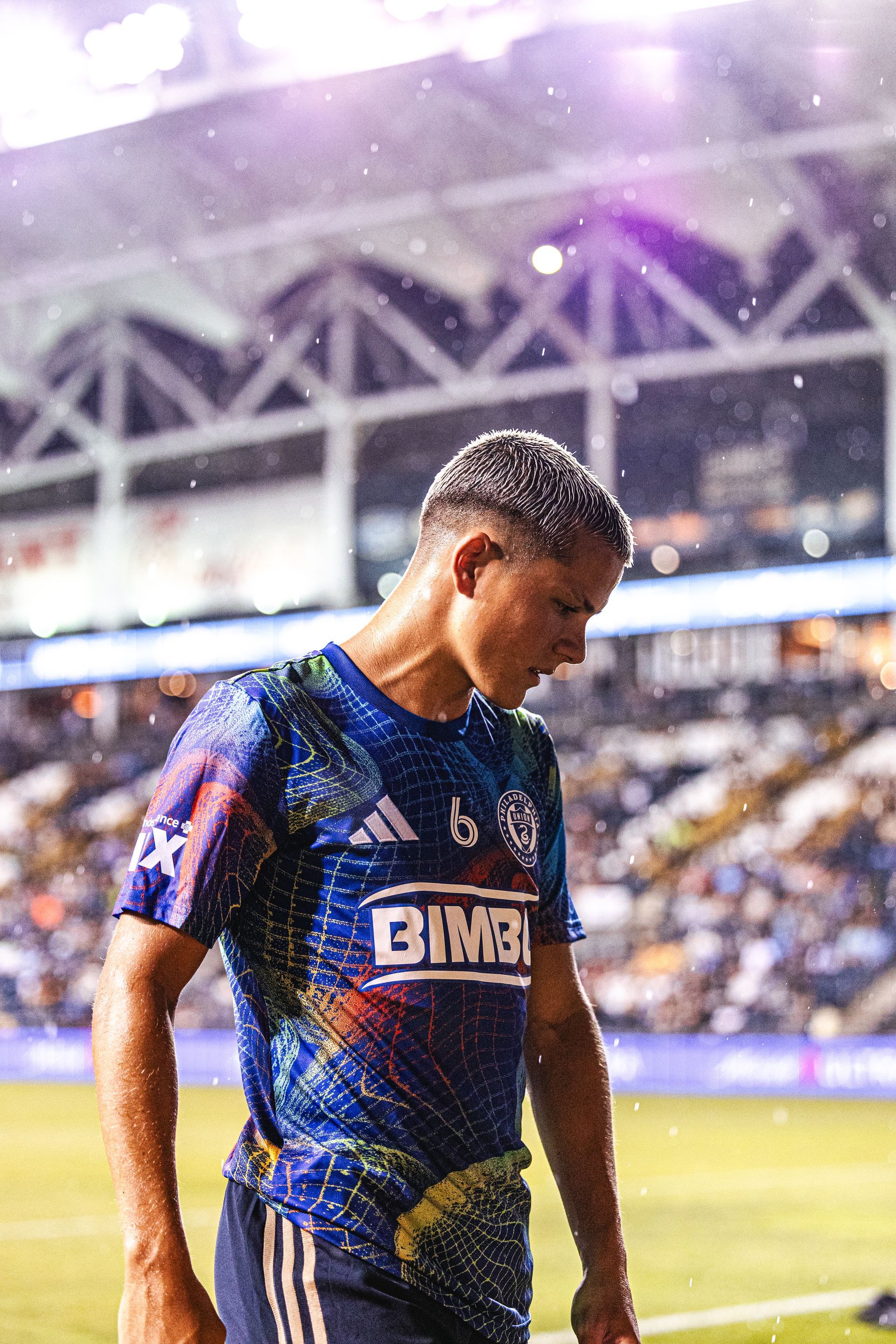 A soccer player in a blue patterned Philadelphia Union jersey stands on the field during a rainstorm at night.