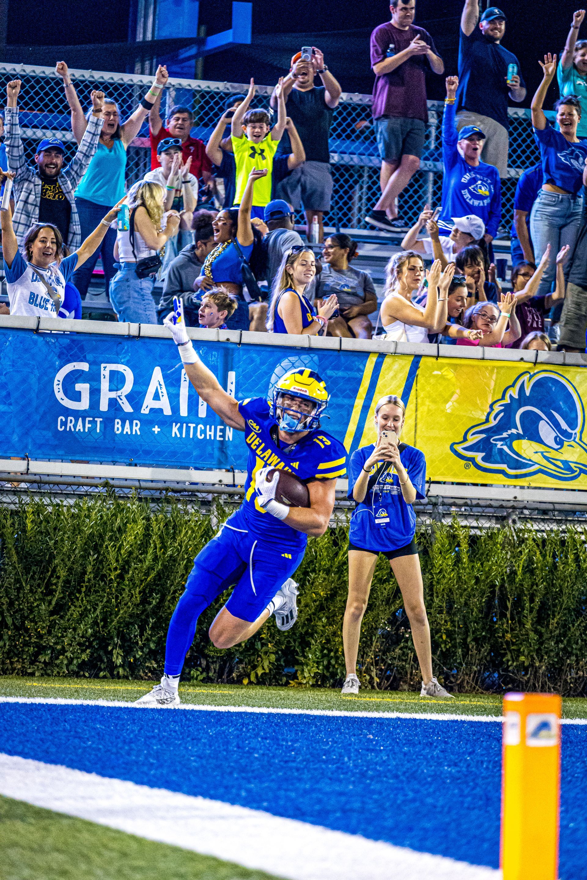 A football player in a blue uniform celebrates a touchdown on the field while fans cheer in the stands behind them.