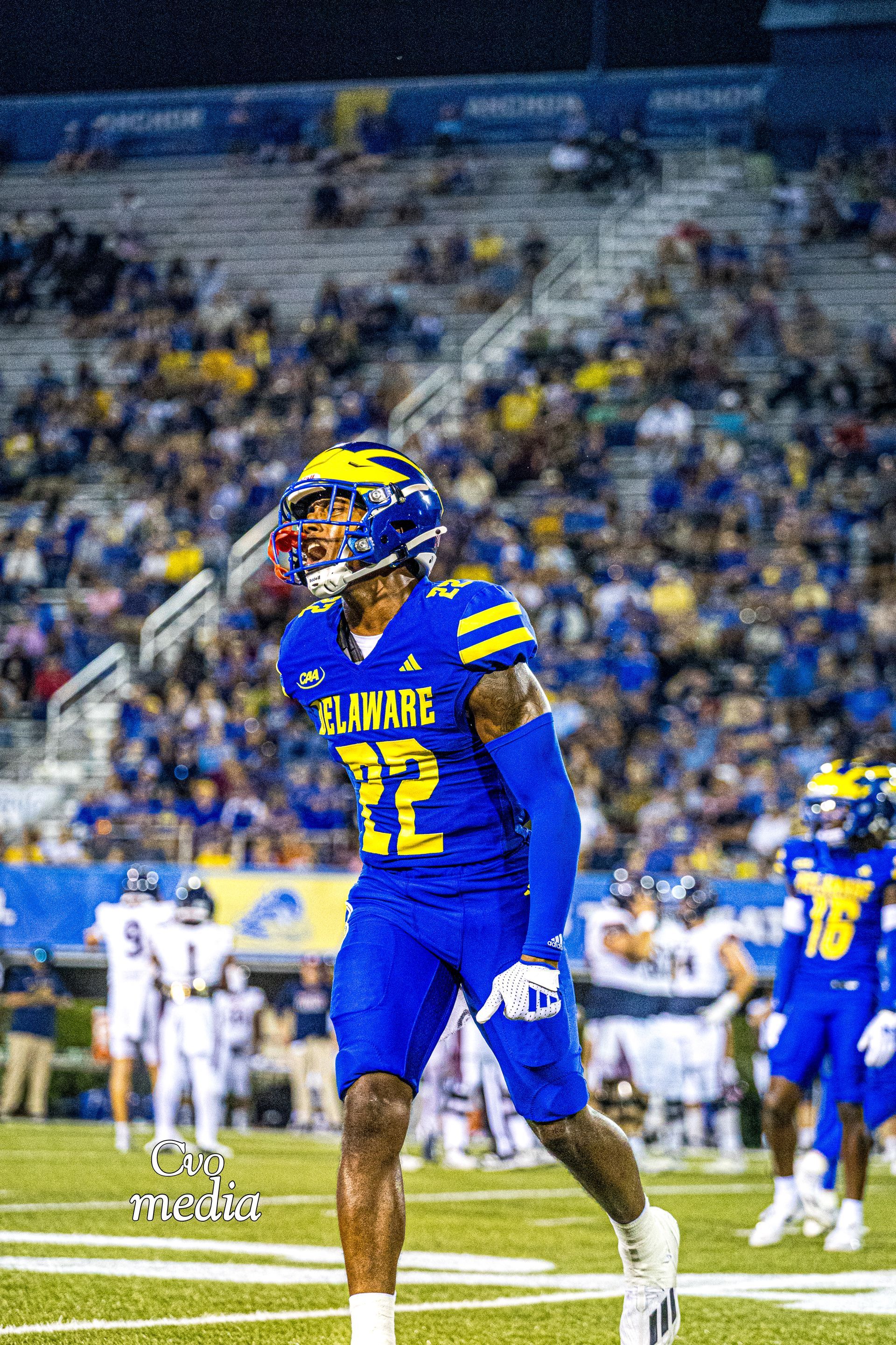 A Delaware football player in blue and yellow gear stands on the field at night with stadium lights in the background.