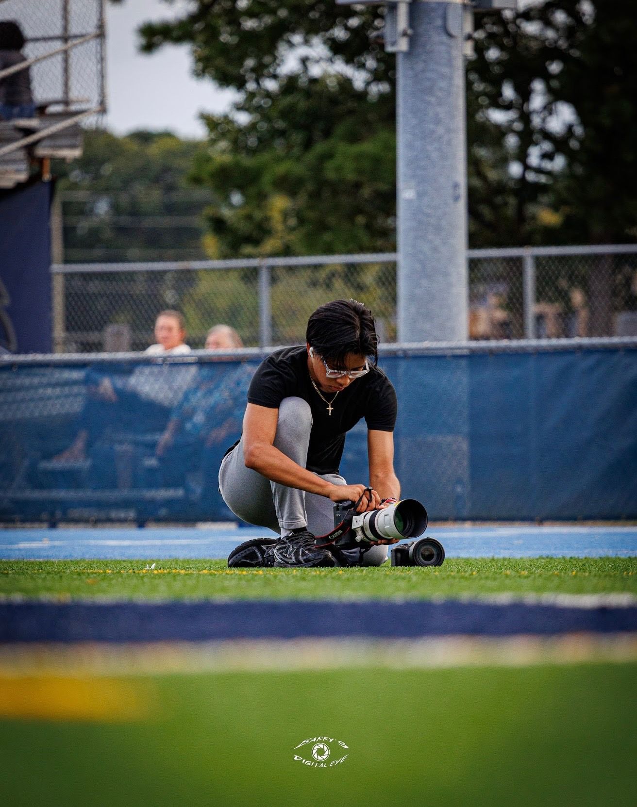 A photographer kneels on a turf field, adjusting a professional camera with a long telephoto lens.