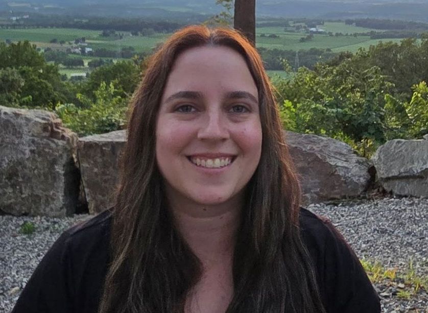 Woman with brown hair smiles at the camera, with a natural landscape background.