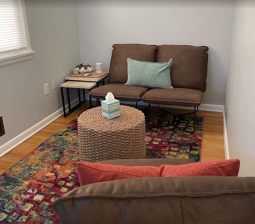 Cozy therapy office with brown loveseat, colorful rug, wicker table, and small side table near a window.