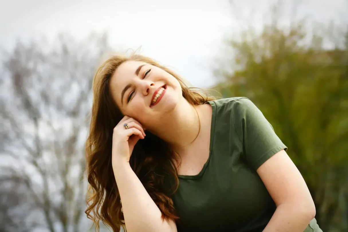 Young woman with brown hair smiles, tilting head, wearing a green shirt outdoors.