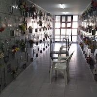 Un pasillo estrecho en un columbario. Paredes grises con nichos que albergan flores. Dos sillas blancas se encuentran cerca de una puerta abierta.