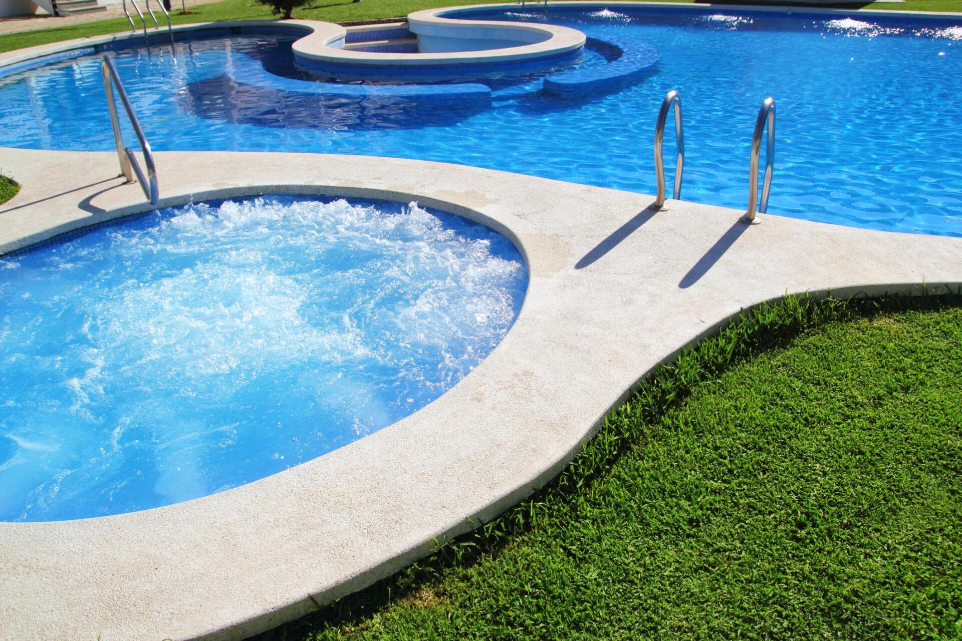 Pool area with a hot tub, steps, and surrounding green grass. Blue water, sunny day.