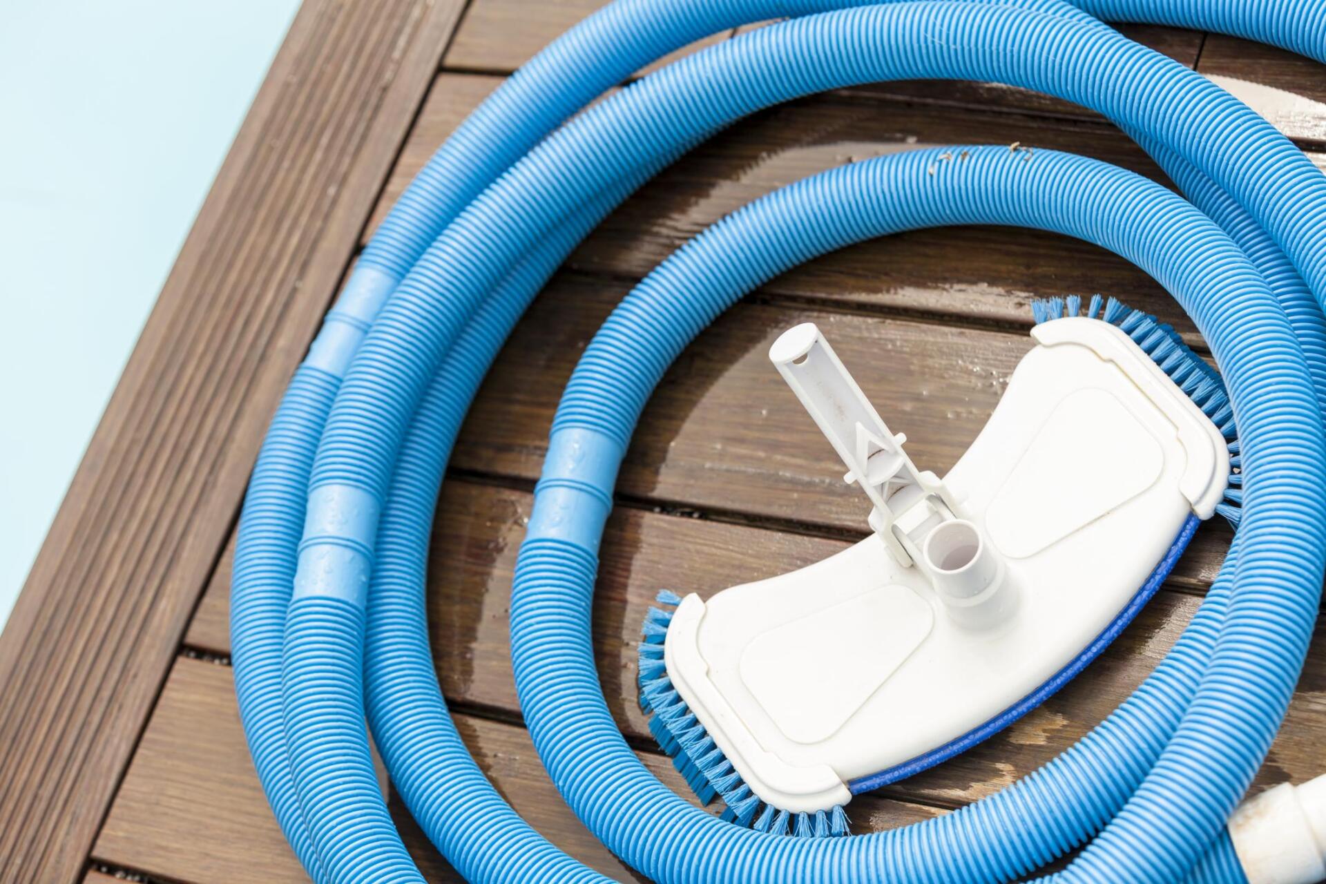 Blue pool vacuum hose and brush coiled on a wooden deck near a pool.