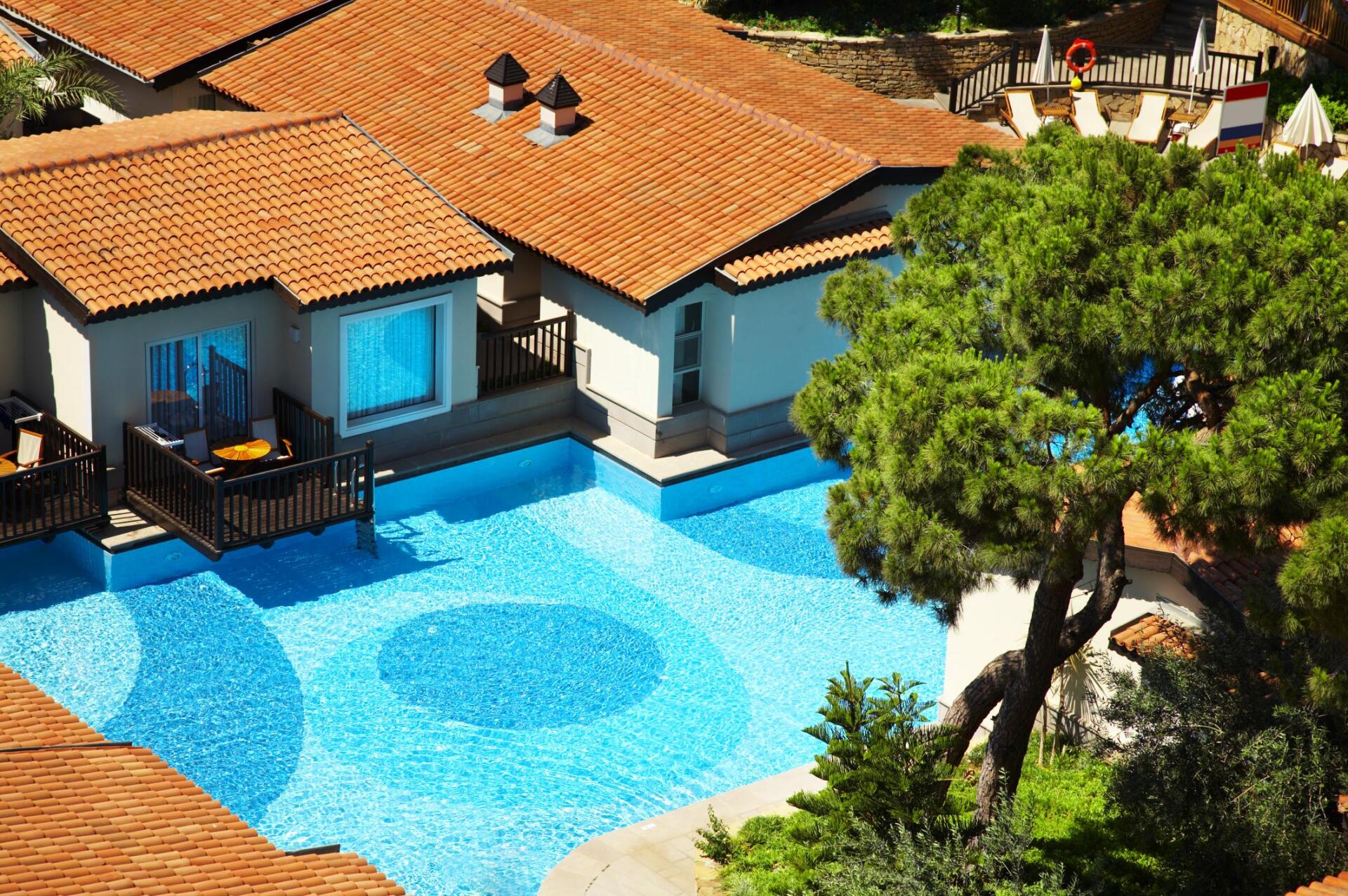 Resort pool with mosaic floor, adjacent buildings with orange tile roofs, sunny day.