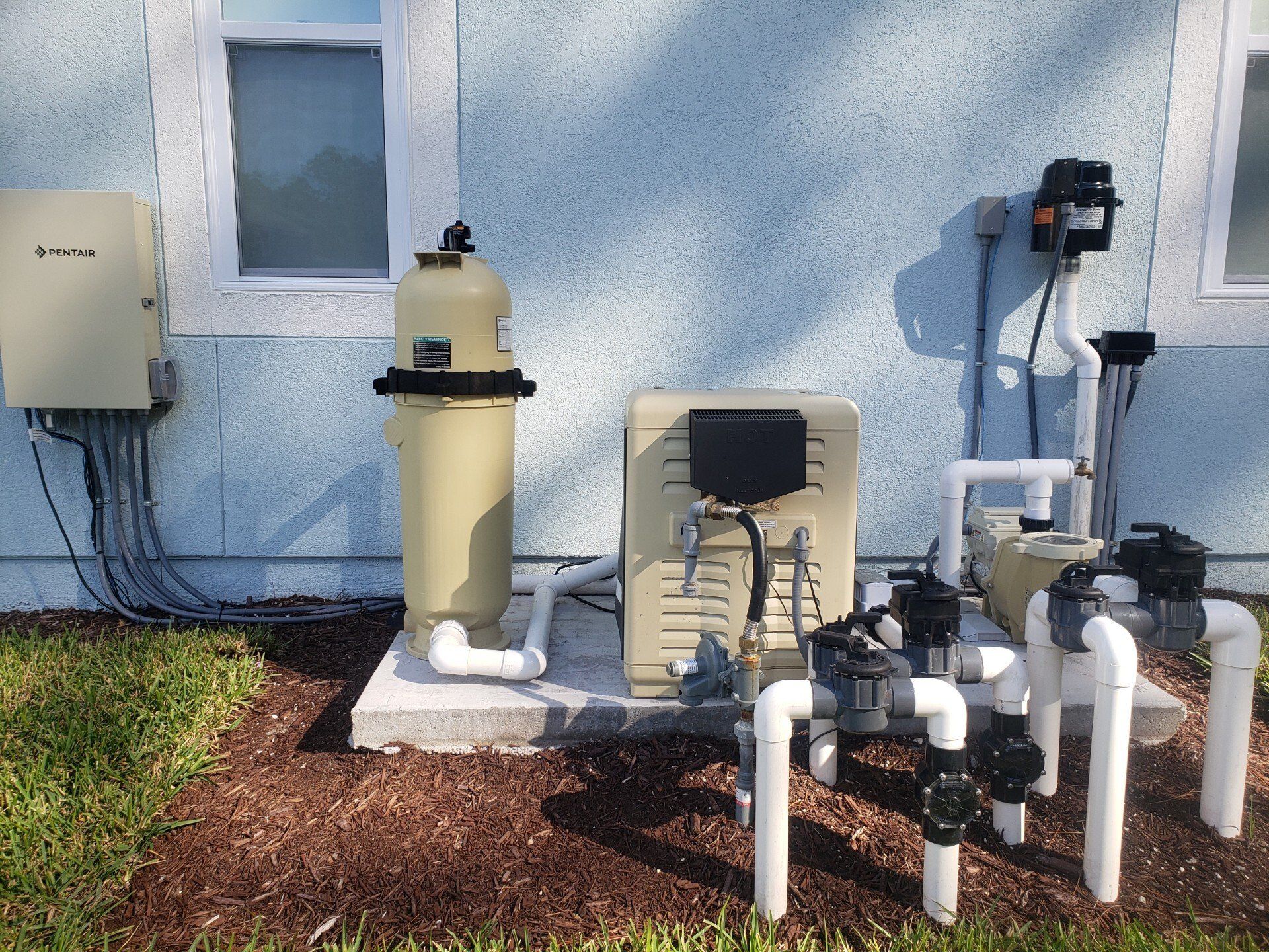 Pool equipment against a light blue wall. Includes a filter, pump, heater, and control box.