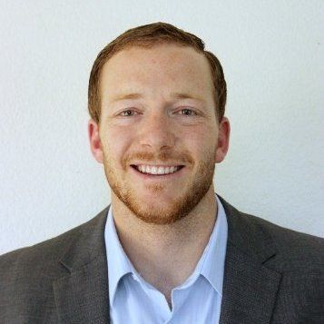Man with red hair and beard smiles, wearing a gray blazer and blue shirt against a white wall.