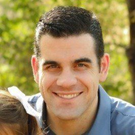 Man with dark hair smiling outdoors, wearing a blue collared shirt.
