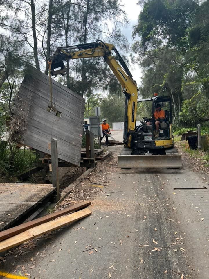 Yellow Excavator Lifting Wooden Barrier on Road. Worker Nearby — ALI Civil Pty Ltd in South Nowra, NSW