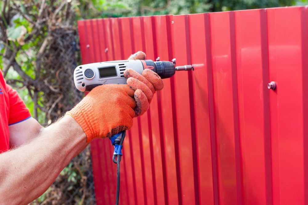 A Man Is Using A Drill To Install A Red Fence — AAA Fencing & Supplies in Southport, QLD