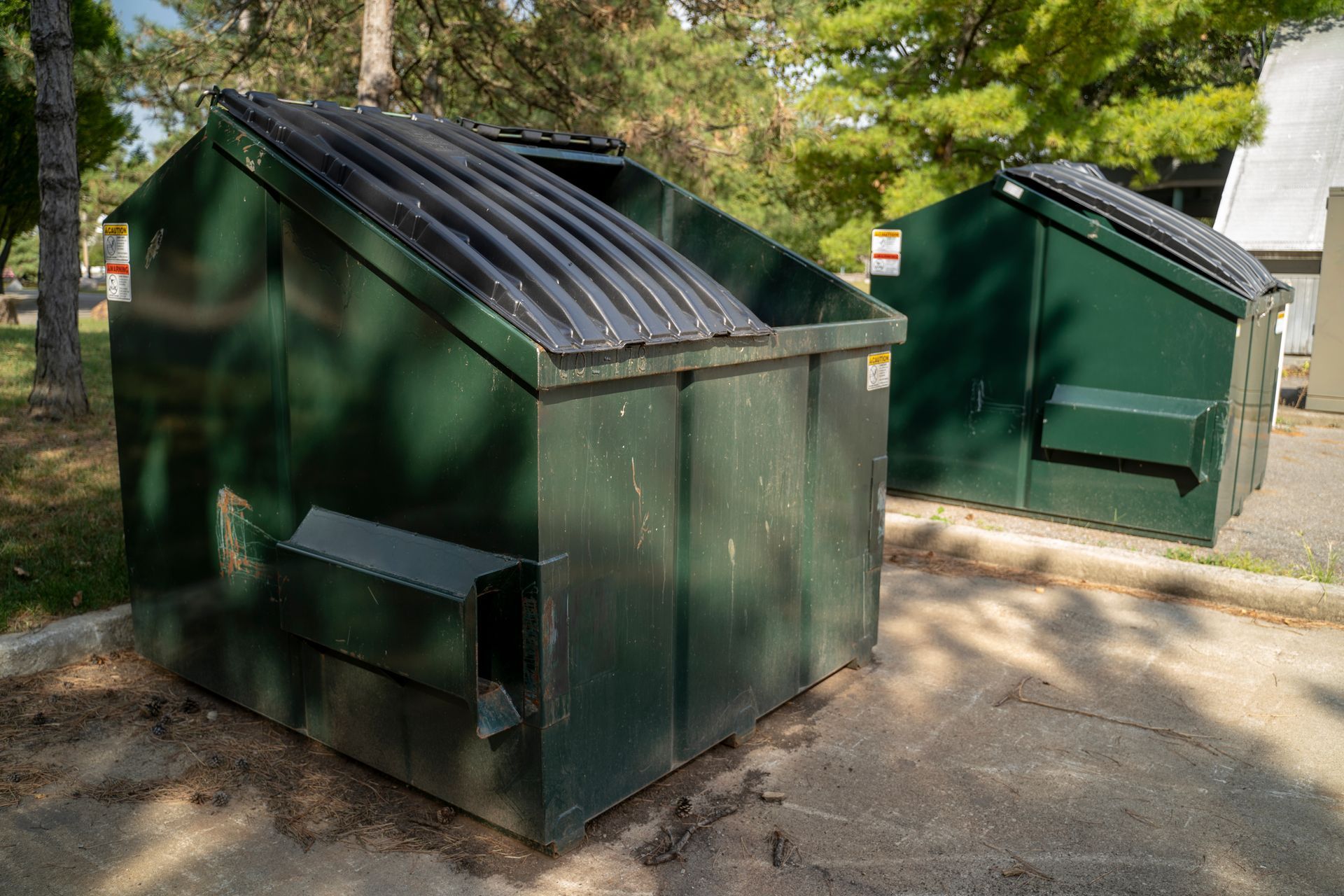 Two green dumpsters on a paved area, with a tree in the background.
