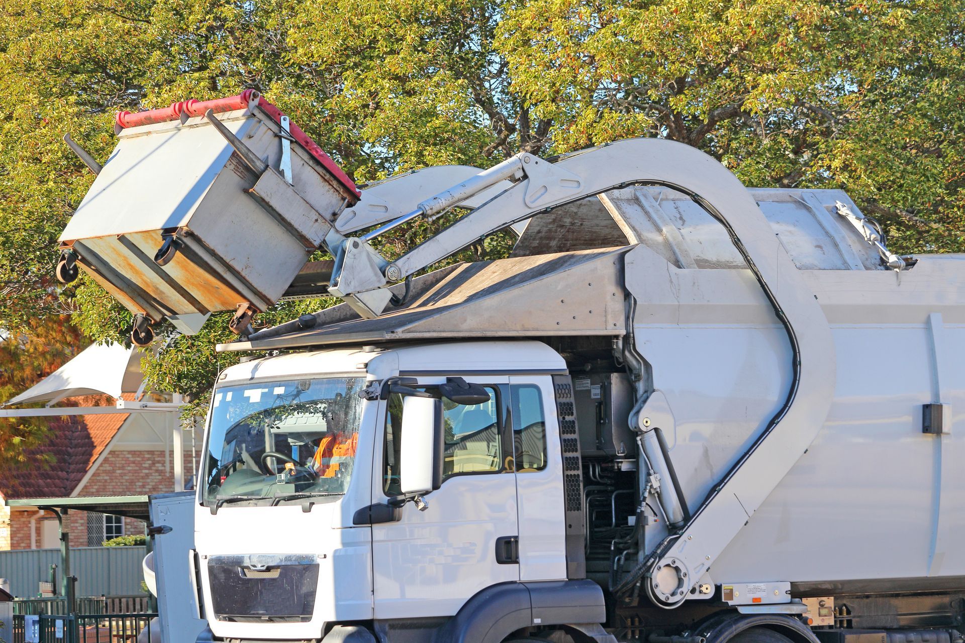 Garbage truck emptying a bin; white truck, red bin, trees in background.