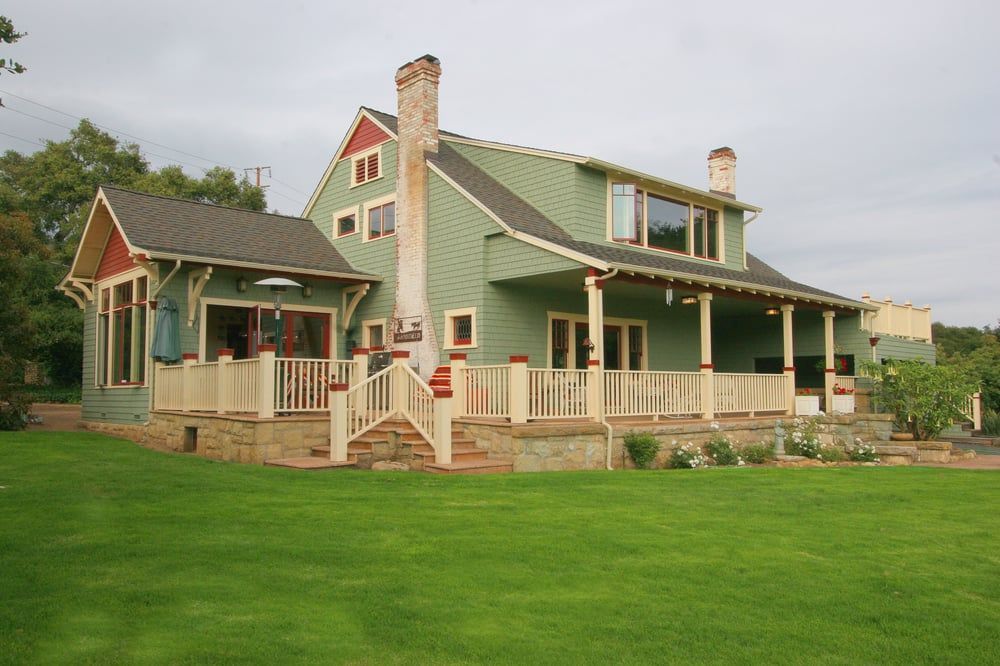 A sage green two-story house with a wrap-around porch, stone base, and chimney, set on a bright green lawn.