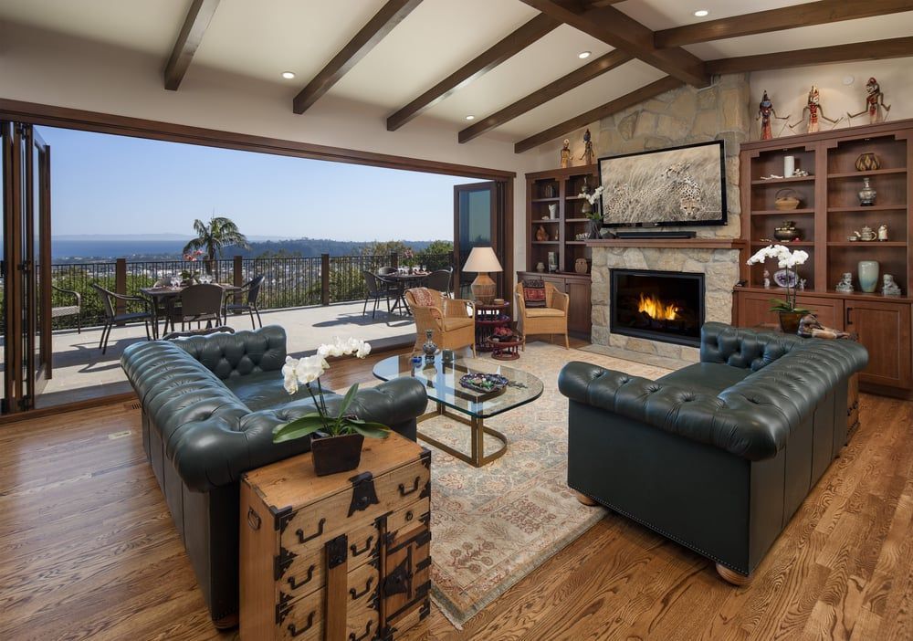 A luxurious living room with dark leather sofas, a stone fireplace, and open glass doors leading to a patio with ocean views.