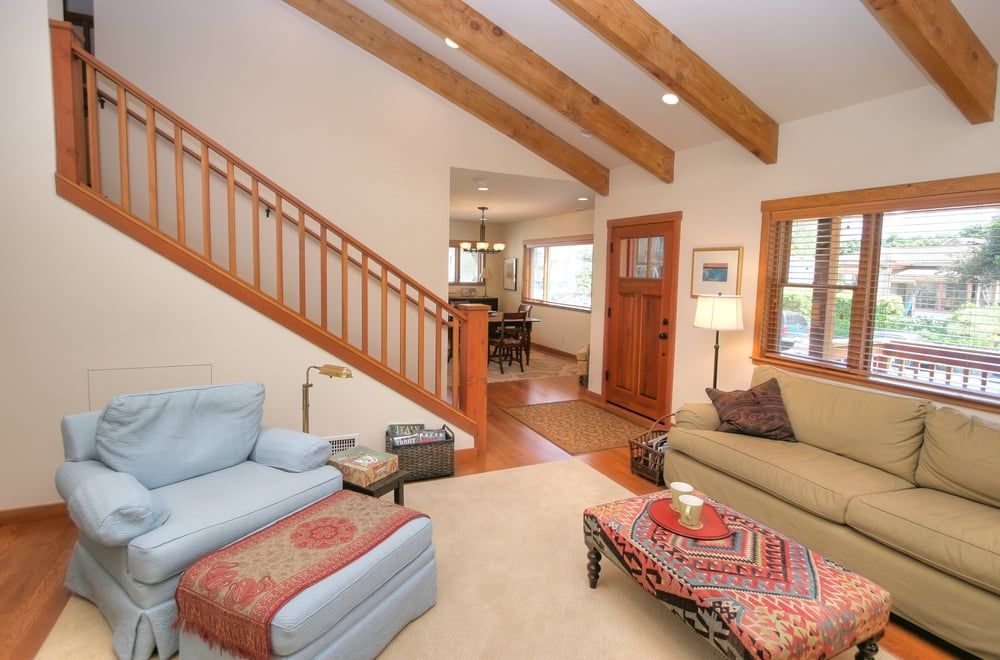 A sunlit living room features a blue armchair, tan sofa with a patterned ottoman, and wooden stairs with beams above.