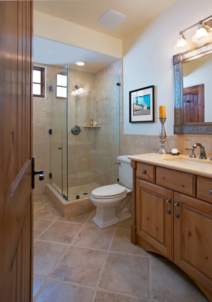 A bright, modern bathroom with a glass-enclosed shower, a white toilet, and a wooden vanity cabinet on stone tile floors.