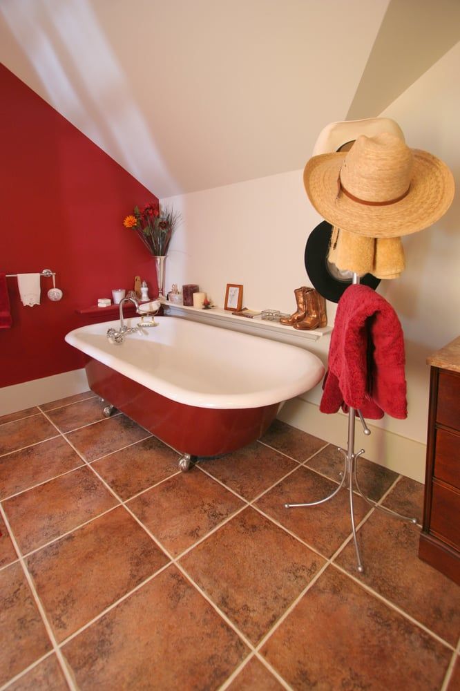 A red clawfoot tub in a bathroom with brown tile floors, a red accent wall, and a hat stand holding hats and a towel.
