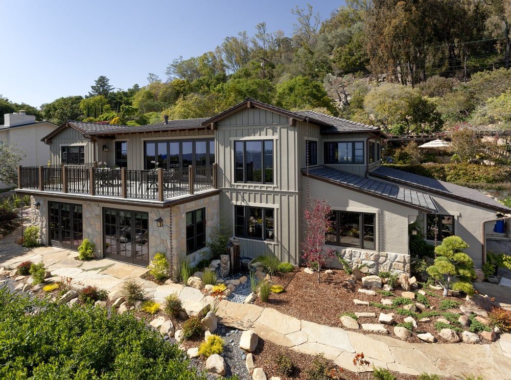 A modern two-story home with neutral siding and stone accents sits on a landscaped, terraced hillside with a large deck.