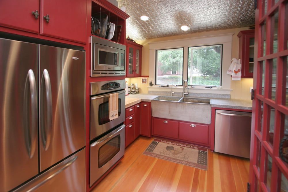 A kitchen with red cabinets, stainless steel appliances, wood floors, and a silver tiled ceiling.