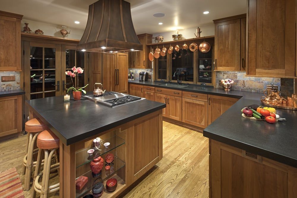 A rustic, wood-cabinet kitchen with a central island, black countertops, a large range hood, and hanging copper cookware.