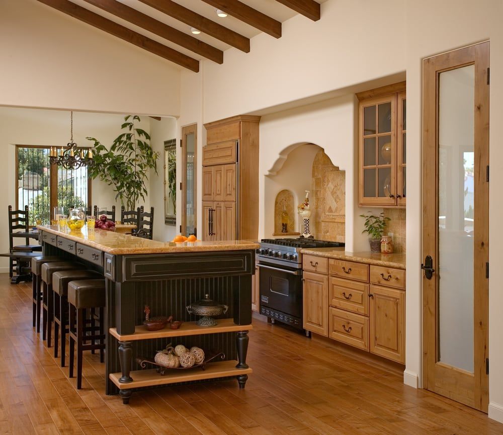 A rustic kitchen featuring a large black island with bar stools, wooden cabinets, and an arched stove alcove.