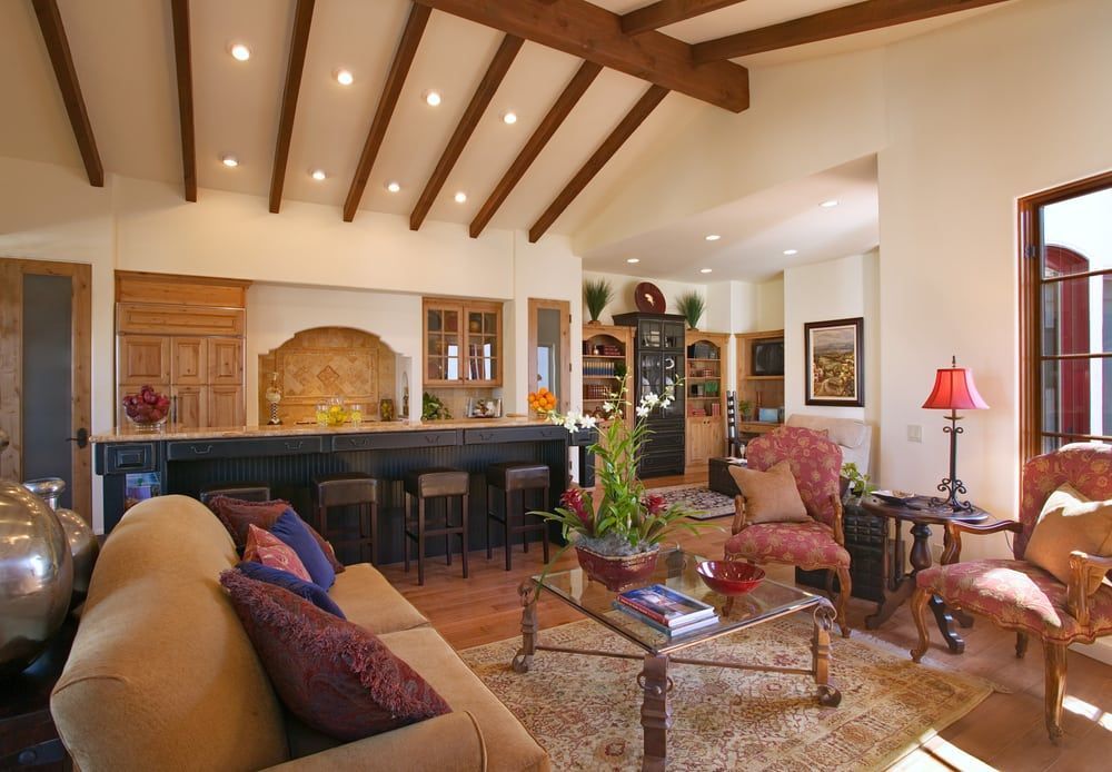 A spacious, warm-toned living room features a vaulted ceiling with wood beams, a breakfast bar, and patterned furniture.