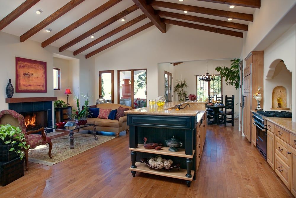 An open-plan living room and kitchen featuring a dark blue island, exposed ceiling beams, fireplace, and hardwood floors.