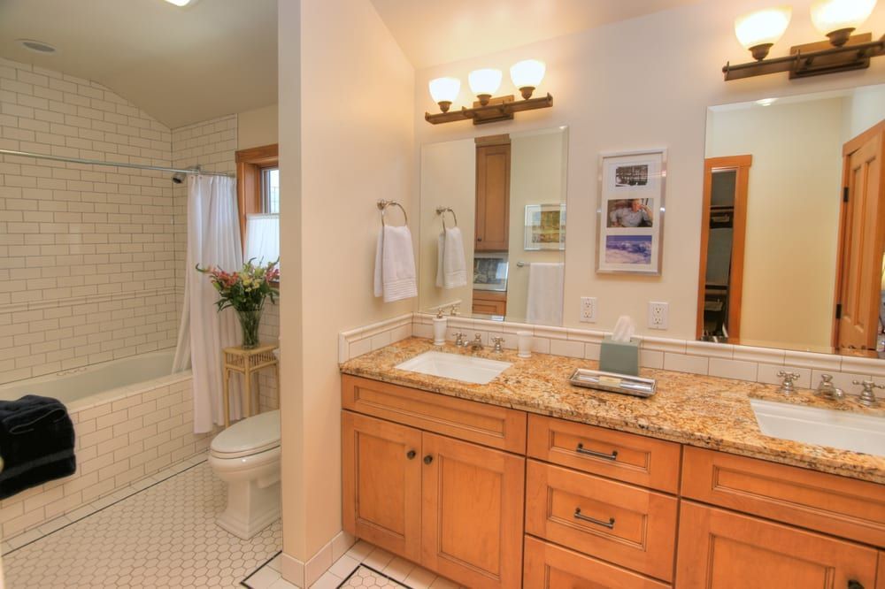 A bathroom with light wood cabinets, granite countertops, dual sinks, a bathtub with white subway tiles, and a toilet.