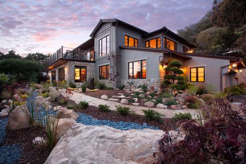 Modern stone house with warm lit windows, landscaped rocks, and a dusk sky