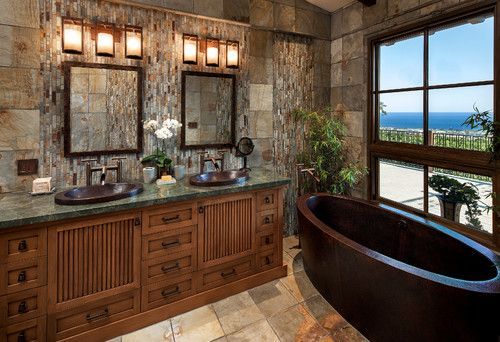 Stone bathroom with wooden vanity, mirrors, and a large black soaking tub by a window overlooking the ocean.