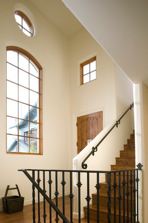Two construction workers wearing yellow hard hats work on the exterior of a wooden house under a clear blue sky.