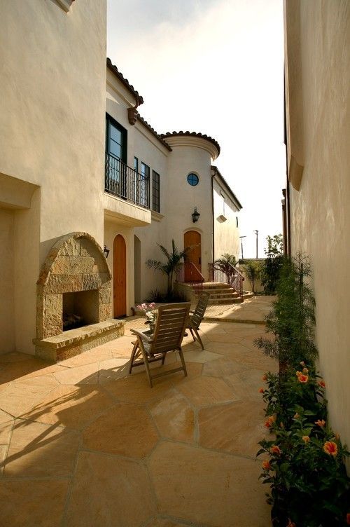 Narrow sunny courtyard walkway between stucco buildings, with potted plants and outdoor chairs.