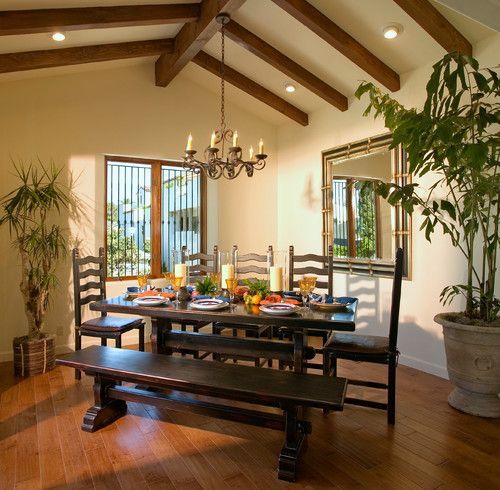 Dining room with a set table, wooden beams, chandelier, and large potted plants near windows