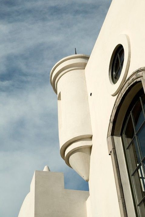 Two construction workers in hard hats work outside a wooden building; one uses a drill on a wooden board.