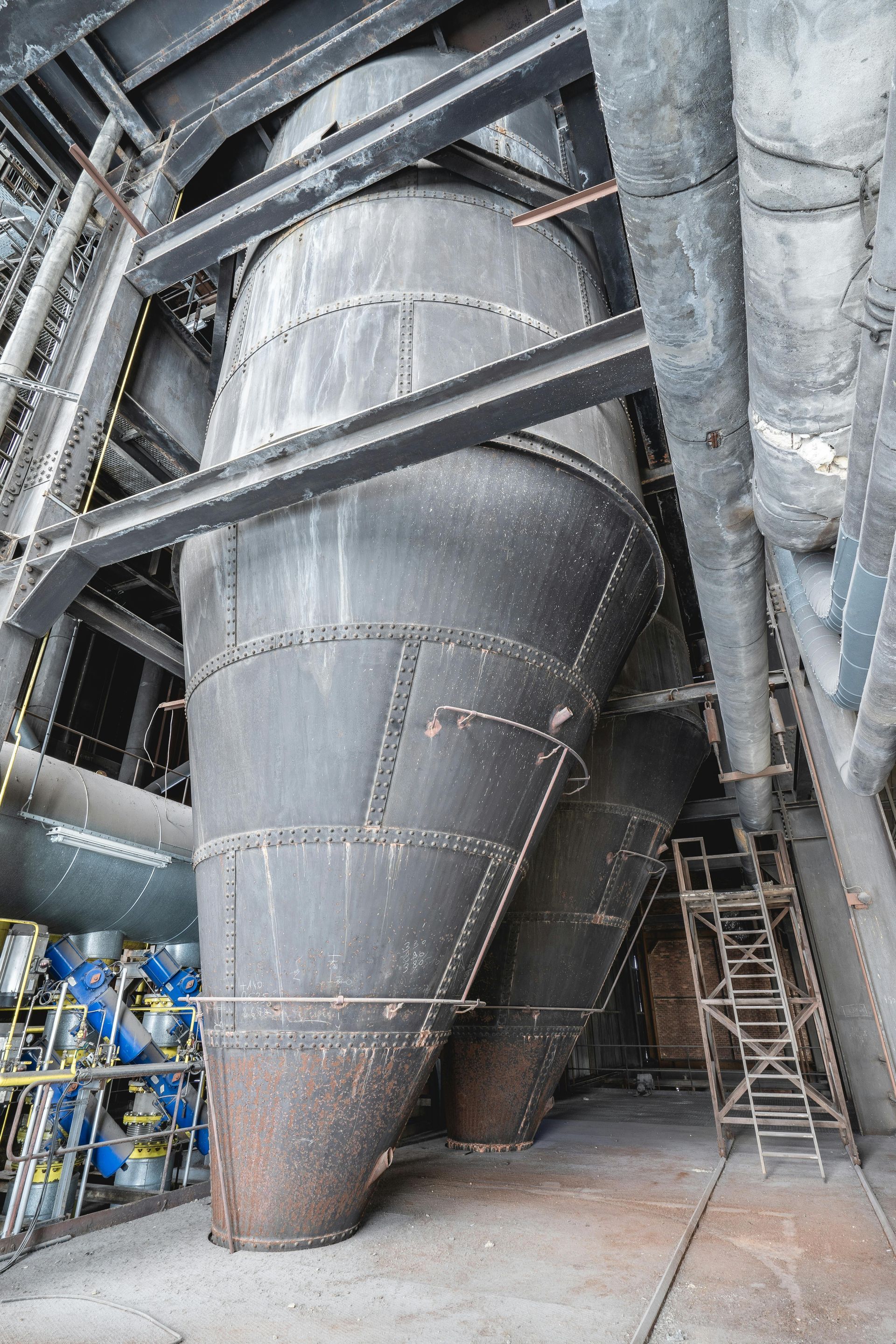 Large, riveted metal industrial hoppers stand in a concrete facility, with pipes and a ladder nearby.