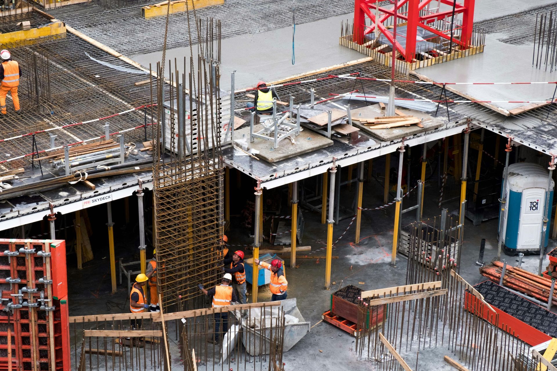 Construction workers in hard hats and high-visibility vests work on a concrete building site with steel rebar and cranes.