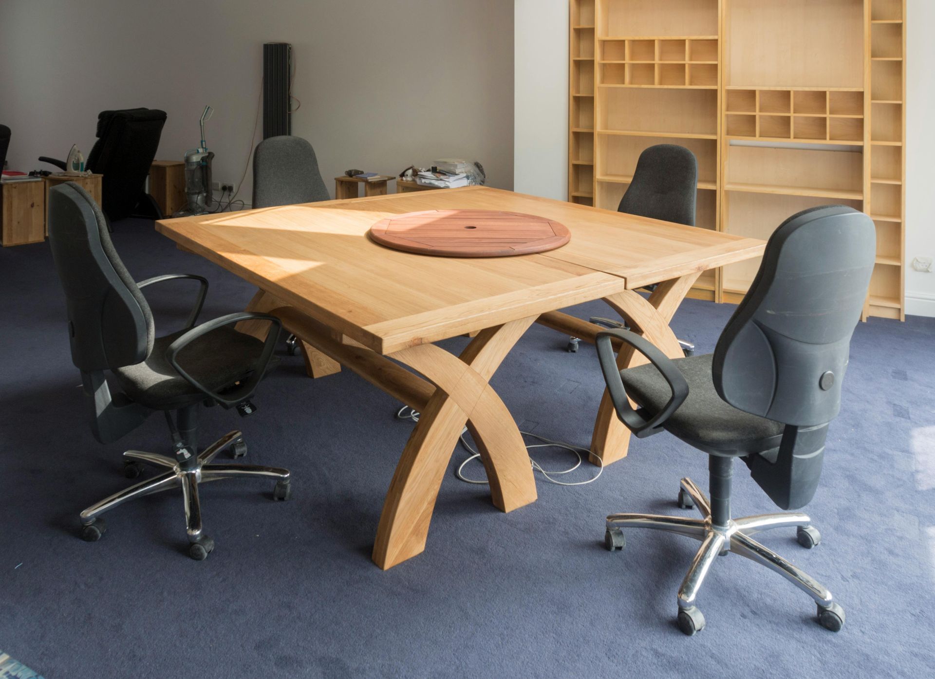 A square wooden meeting table with an X-shaped base and four black rolling chairs on a blue carpeted floor.