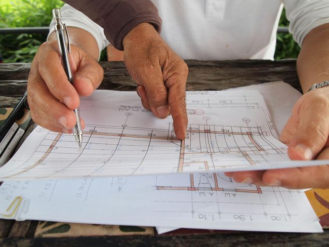 Hands pointing to and writing on architectural floor plans on a wooden table.
